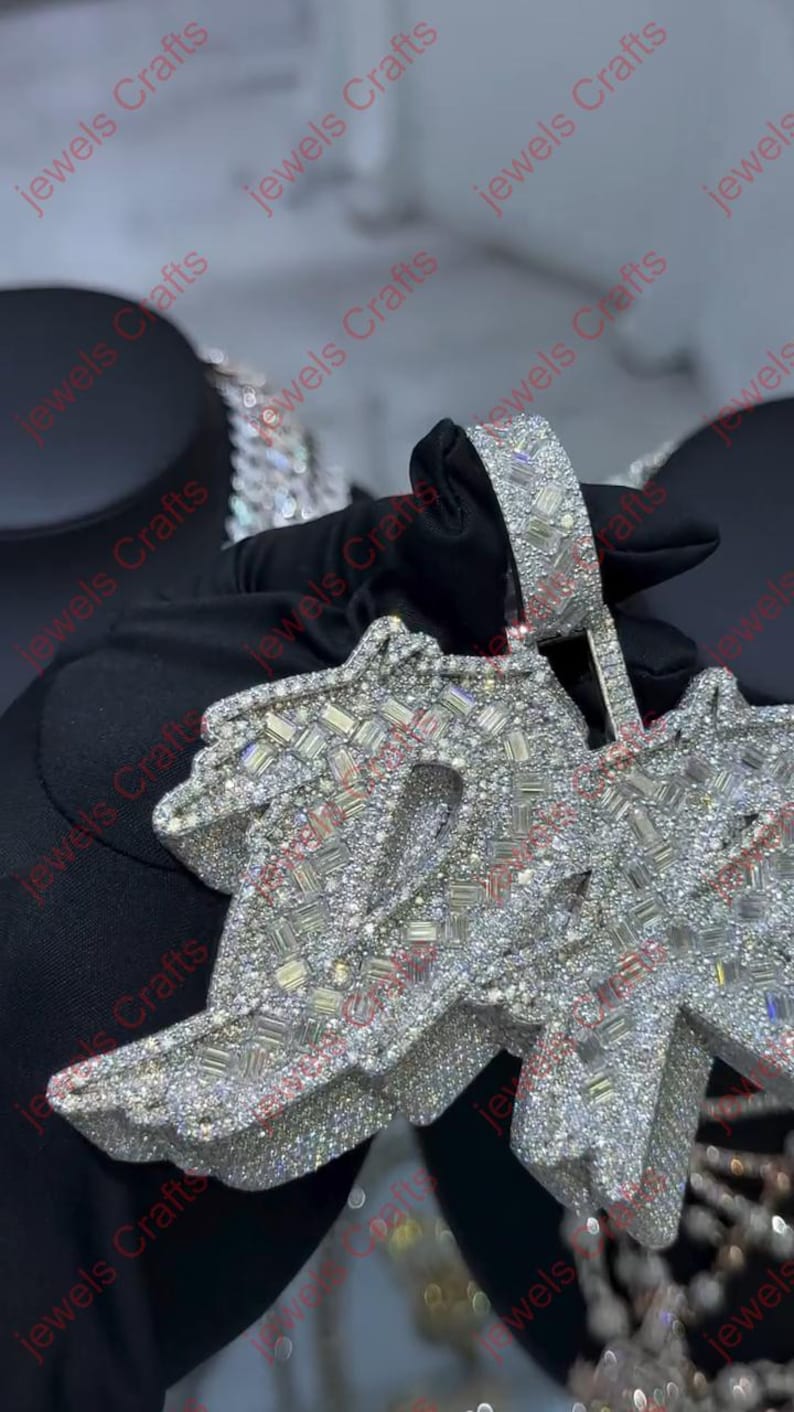 May include: A large, silver-toned pendant with a stylized design, heavily encrusted with sparkling, clear gemstones. The pendant is held by a gloved hand, showcasing the intricate details and craftsmanship of the jewelry.