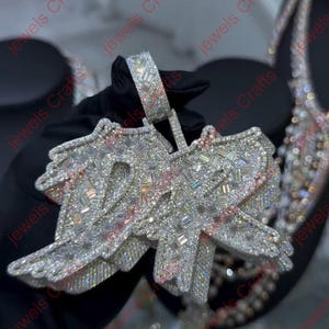 May include: A large, sparkling pendant with the letters "DR" encrusted with numerous clear gemstones. The pendant has a silver-colored metal base with pink accents. The bail is also encrusted with gemstones.
