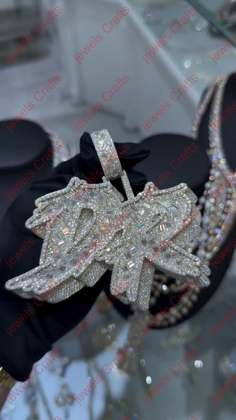May include: A large, sparkling pendant with the letters "DR" encrusted with numerous clear gemstones. The pendant has a silver-colored metal base with pink accents. The bail is also encrusted with gemstones.