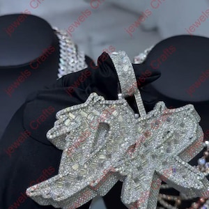 May include: A diamond-encrusted pendant in the shape of the word "Jewels". The pendant is silver-colored and features numerous small, sparkling stones. The background is dark, highlighting the jewelry's brilliance. The pendant is held by a gloved hand.