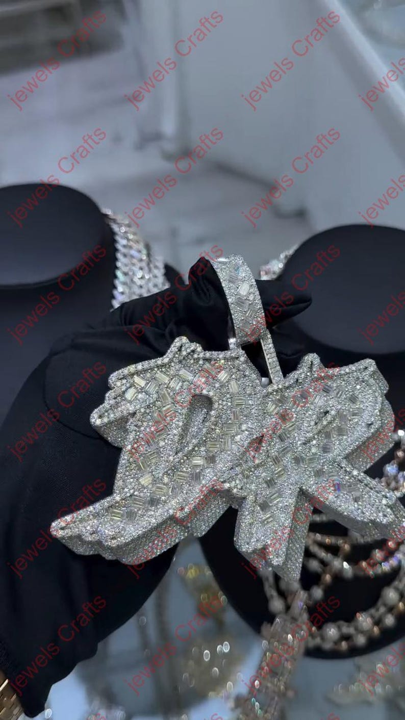 May include: A diamond-encrusted pendant in the shape of the word "Jewels". The pendant is silver-colored and features numerous small, sparkling stones. The background is dark, highlighting the jewelry's brilliance. The pendant is held by a gloved hand.