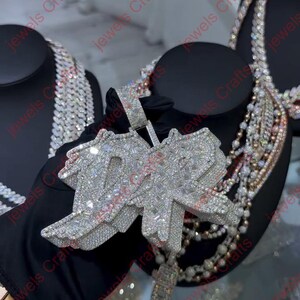 May include: A diamond-encrusted pendant with the letters "DR" in a stylized design. The pendant is held by a gloved hand, with other jewelry pieces visible in the background. The pendant has a rectangular bale and a dangling rectangular charm.