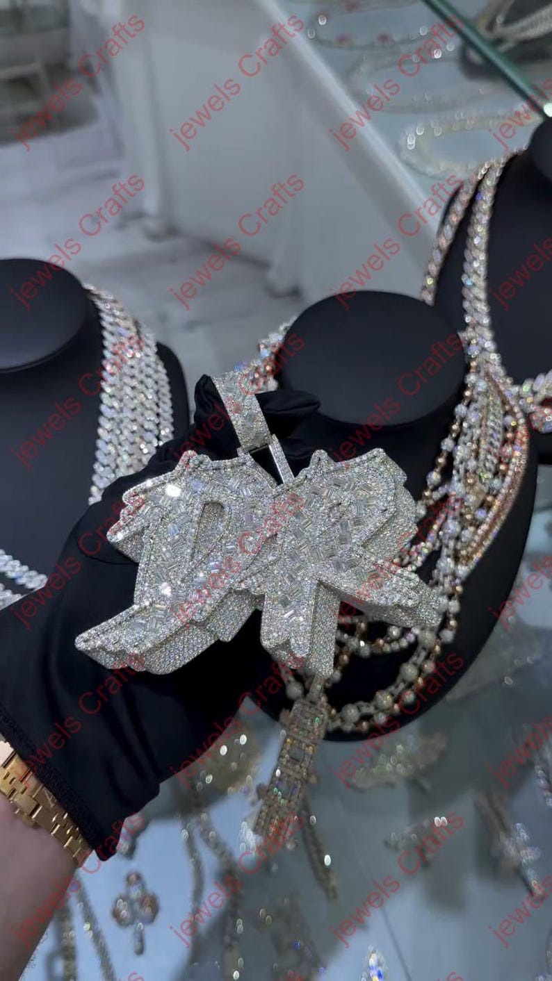 May include: A diamond-encrusted pendant with the letters "DR" in a stylized design. The pendant is held by a gloved hand, with other jewelry pieces visible in the background. The pendant has a rectangular bale and a dangling rectangular charm.
