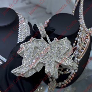 May include: A large, silver-toned pendant with the letters "10" encrusted with numerous small, sparkling stones. The pendant is held by a gloved hand, with other jewelry visible in the background.