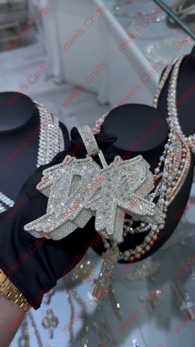 May include: A large, silver-toned pendant with the letters "10" encrusted with numerous small, sparkling stones. The pendant is held by a gloved hand, with other jewelry visible in the background.