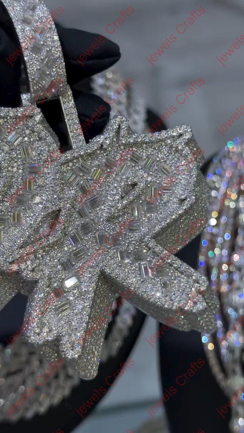 May include: Close-up of a diamond-encrusted pendant. The pendant is shaped like a stylized letter and is covered in small, sparkling diamonds. The background is blurred, highlighting the jewelry.