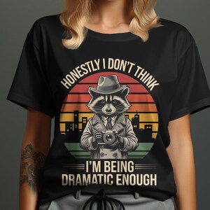 Puede incluir: Camiseta negra con un mapache con sombrero y cámara, sobre un fondo de puesta de sol y horizonte urbano. El texto dice: "HONESTAMENTE, NO CREO QUE ESTÉ SIENDO LO SUFICIENTEMENTE DRAMÁTICO."