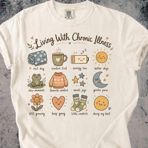 Może przedstawiać: Kremowy t-shirt z napisem Living With Chronic Illness oraz dwunastoma ilustracjami z podpisami: koc, kubek, rozładowana bateria, słońce, żaba, sweter, gwiazdy, księżyc, kwiat, serce, skarpetki i uśmiechnięta buźka.