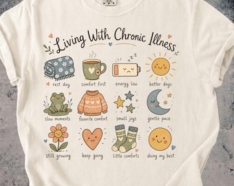 Może przedstawiać: Kremowy t-shirt z napisem Living With Chronic Illness oraz dwunastoma ilustracjami z podpisami: koc, kubek, rozładowana bateria, słońce, żaba, sweter, gwiazdy, księżyc, kwiat, serce, skarpetki i uśmiechnięta buźka.