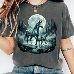 以下が含まれることがあります： 暗い灰色のTシャツ。霧のかかった風景の中に3頭の馬が描かれた穏やかなイラスト。満月、山、木々が描かれ、穏やかなシーンを演出。馬はグレーとブルーの色合いで表現されています。