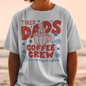 Op de afbeelding: Een lichtgrijs t-shirt met een grafisch ontwerp met de rode tekst TIRED DADS en COFFEE CREW. Een centrale illustratie toont een dampende koffiemok omringd door blauwe sterren, bloemen en de kleine blauwe tekst running on caffeine.