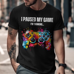 Könnte beinhalten: Schwarzes T-Shirt mit dem Text "I PAUSED MY GAME I'M THINKING..." und einem bunten, abstrakten Design eines Videospiel-Controllers. Der Controller hat verschiedene Farben und Knöpfe.