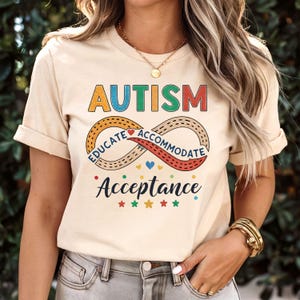 Puede incluir: Camiseta beige con la palabra "AUTISMO" en letras coloridas. Debajo, un diseño de símbolo de infinito con las palabras "EDUCAR" y "ACOMODAR" y la palabra "ACEPTACIÓN". Estrellas y corazones coloridos completan el diseño.