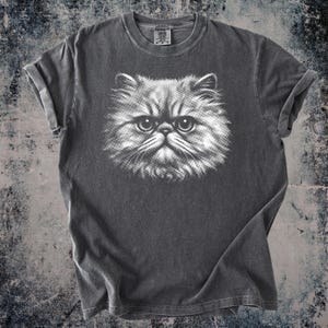 May include: A charcoal gray t-shirt featuring a detailed white illustration of a Persian cat's face. The cat's eyes are large and expressive, with a fluffy, textured appearance. The sleeves are rolled up.