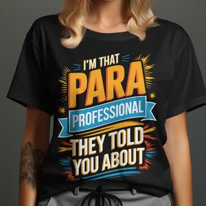 以下が含まれることがあります： 黒いTシャツ。オレンジ、青、黄色の文字で「I'M THAT PARA PROFESSIONAL THEY TOLD YOU ABOUT」と書かれています。テキストは太字で様式化されています。半袖のシャツです。