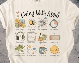 May include: An off-white t-shirt featuring the text Living With ADHD and a grid of twelve colorful illustrations representing various experiences, including a bee, a clock, headphones, a to-do list, a frog, and a coffee mug with descriptive labels.