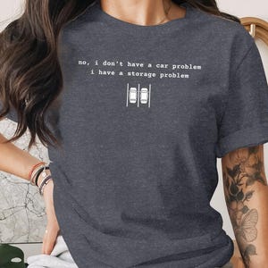 Puede incluir: Camiseta gris oscuro con el texto "no, I don't have a car problem I have a storage problem." La camiseta presenta un gráfico de dos coches aparcados en una plaza de aparcamiento. Una camiseta informal para el día a día.