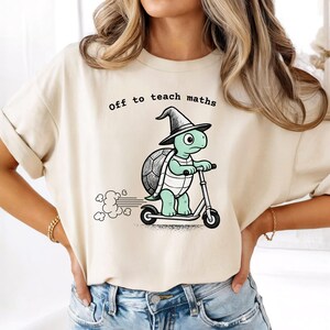 Op de afbeelding: Beige T-shirt met de tekst "off to teach maths". Het shirt heeft een cartoon schildpad met een heksenhoed op een scooter. De schildpad is lichtgroen met een donkergroene schild.