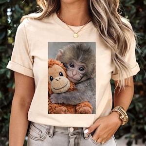 Op de afbeelding: Beige T-shirt met een afbeelding van een baby aap die een bruine knuffel aap omhelst. De baby aap heeft grijs haar en grote ogen. Het knuffel heeft een lachend gezicht.