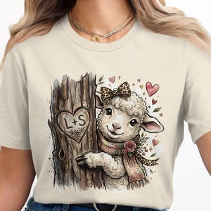 Op de afbeelding: Crèmekleurig T-shirt met een aquarel illustratie van een schaap met een luipaardprint strik en sjaal, en bloemaccenten. Het schaap staat naast een boomstam met een hart waarin de letters "L+S" zijn gegraveerd.