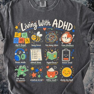 Può includere: Una t-shirt grigio antracite con il testo Living With ADHD e una griglia di illustrazioni colorate che rappresentano esperienze quotidiane, tra cui post-it, un'ape, un timer, cuffie, una lista di cose da fare, un libro, una batteria, una rana e una tazza di caffè.