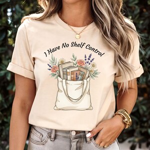 Puede incluir: Camiseta beige con el texto "I Have No Shelf Control" sobre un dibujo de una bolsa de mano llena de libros y flores. La bolsa es blanca y las flores incluyen rosas, margaritas y lavanda.