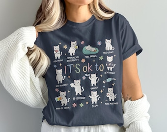 May include: Navy blue t-shirt with a graphic featuring cartoon cats and the text "IT'S ok to" followed by various affirmations. The cats are depicted in different poses, each representing a positive action or feeling.