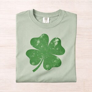 May include: A light green t-shirt featuring a large, distressed green four-leaf clover graphic. The shirt is folded neatly, showcasing the design prominently. The shirt is made of a soft material and is perfect for St. Patrick's Day.