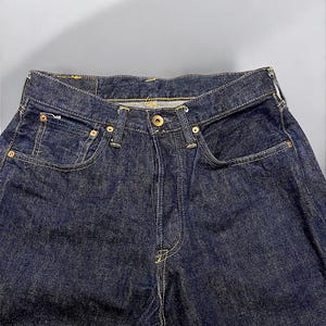 K&ouml;nnte beinhalten: Nahaufnahme einer dunkelblauen Jeans. Die Jeans hat einen Knopfverschluss, Vordertaschen und eine M&uuml;nztasche. Die N&auml;hte sind gelb und die Nieten goldfarben. Die Jeans ist ein klassischer Stil.