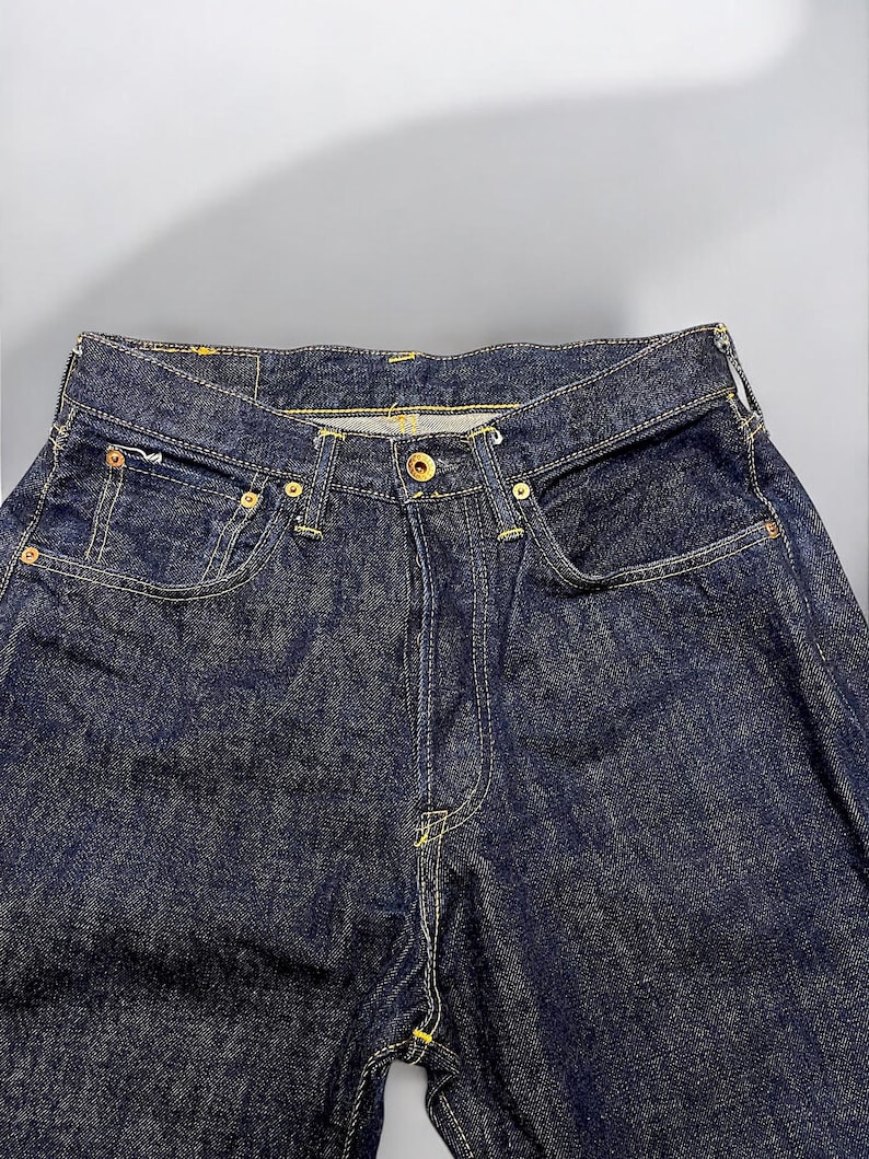 K&ouml;nnte beinhalten: Nahaufnahme einer dunkelblauen Jeans. Die Jeans hat einen Knopfverschluss, Vordertaschen und eine M&uuml;nztasche. Die N&auml;hte sind gelb und die Nieten goldfarben. Die Jeans ist ein klassischer Stil.