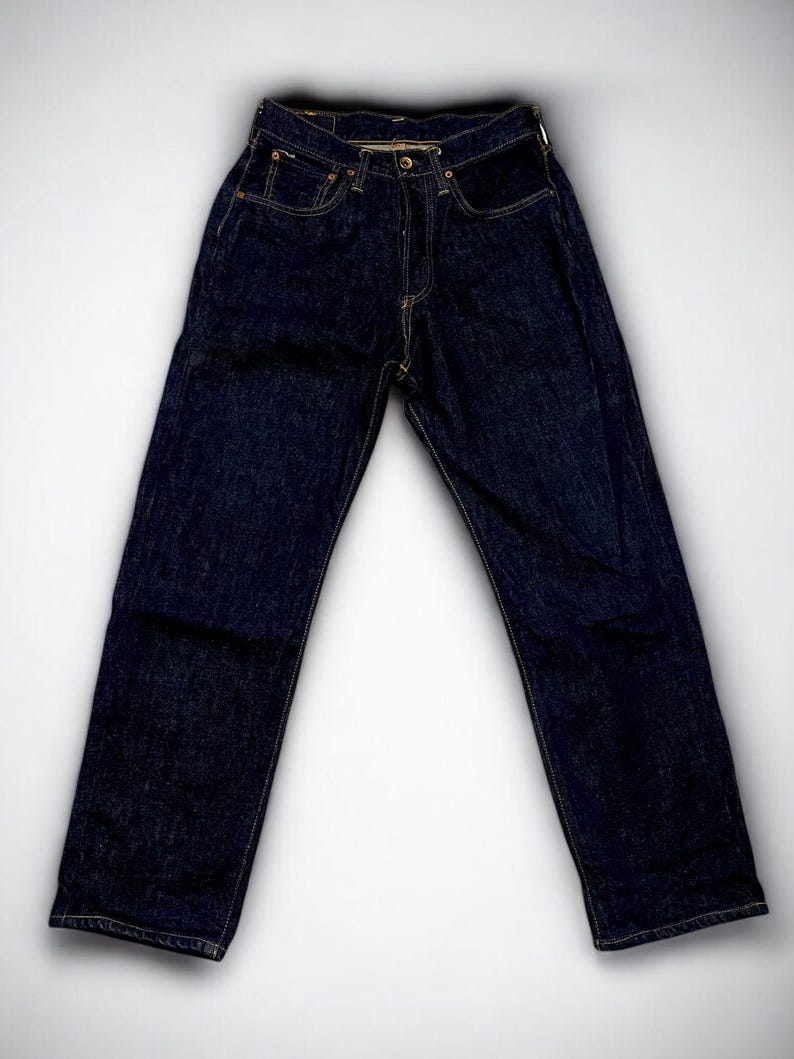 K&ouml;nnte beinhalten: Dunkelblaue Jeans mit klassischem F&uuml;nf-Taschen-Design. Die Jeans hat einen Knopf- und Rei&szlig;verschluss und sichtbare N&auml;hte in kontrastierendem gelbem Faden. Die Jeans liegen flach auf einem wei&szlig;en Hintergrund.