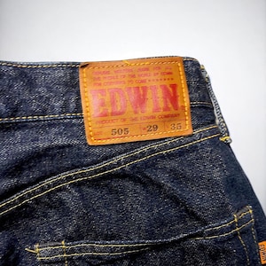 K&ouml;nnte beinhalten: Nahaufnahme einer dunkelblauen Jeans mit gelben N&auml;hten. Ein braunes Lederetikett mit dem Markennamen "EDWIN" in roter Schrift ist sichtbar. Das Etikett enth&auml;lt Text und Zahlen. Die Jeans ist ein klassischer Stil.