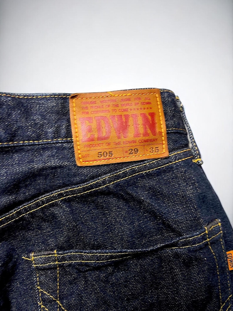 K&ouml;nnte beinhalten: Nahaufnahme einer dunkelblauen Jeans mit gelben N&auml;hten. Ein braunes Lederetikett mit dem Markennamen "EDWIN" in roter Schrift ist sichtbar. Das Etikett enth&auml;lt Text und Zahlen. Die Jeans ist ein klassischer Stil.