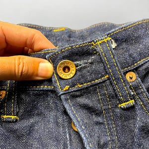 K&ouml;nnte beinhalten: Nahaufnahme einer dunkelblauen Jeans mit gelben N&auml;hten. Die Jeans hat einen Knopfverschluss mit dem Markennamen "EDWIN" auf dem Knopf. Das Bild zeigt die Textur und die Details des Stoffes und der N&auml;hte.