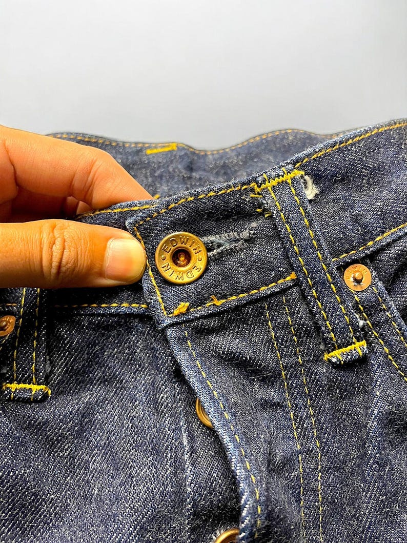 K&ouml;nnte beinhalten: Nahaufnahme einer dunkelblauen Jeans mit gelben N&auml;hten. Die Jeans hat einen Knopfverschluss mit dem Markennamen "EDWIN" auf dem Knopf. Das Bild zeigt die Textur und die Details des Stoffes und der N&auml;hte.