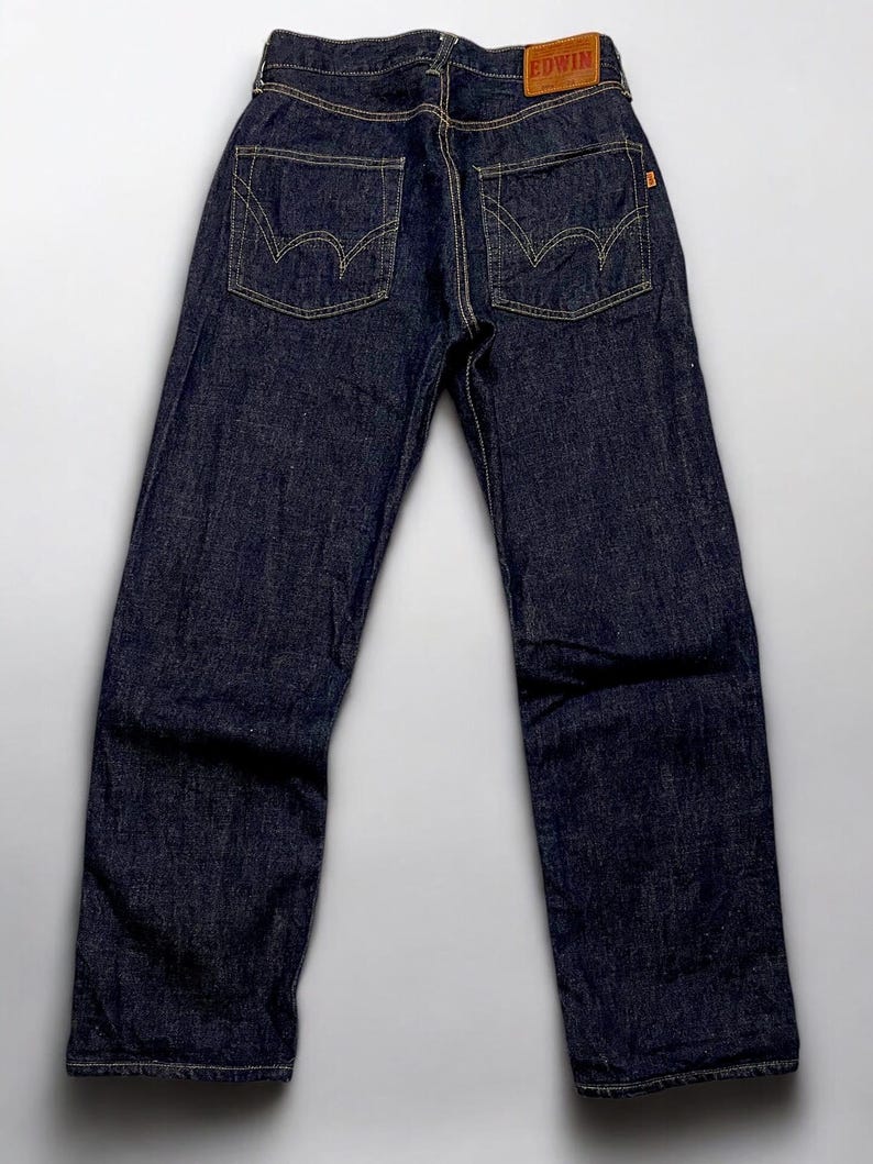 K&ouml;nnte beinhalten: Dunkelblaue Jeans mit gelben N&auml;hten. Die R&uuml;ckseite der Jeans hat zwei Taschen mit einem einzigartigen geschwungenen Design. Ein braunes Lederetikett mit dem Markennamen ist am Bund sichtbar. Die Jeans haben eine klassische, gerade Passform.