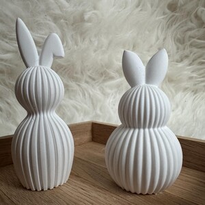 May include: Two white ceramic bunny ornaments with ribbed bodies and long ears. The bunnies are displayed on a wooden tray against a fluffy white background. The ornaments have a minimalist, modern design, suitable for Easter decorations.