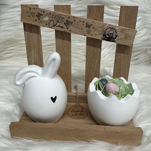 May include: An Easter-themed wooden display. It features a white ceramic bunny with a black heart, a white eggshell-shaped container with eggs, and a sign reading "Frohe Ostern". The display is handmade with love.