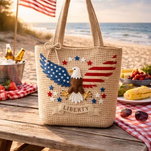 May include: A beige tote bag with an American flag and eagle design. The bag features blue and red stars and stripes, with the word "LIBERTY" on a banner. The bag is on a wooden picnic table near the beach.