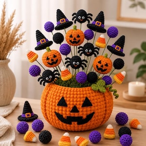 May include: A crocheted Halloween centerpiece featuring a large orange jack-o'-lantern bowl filled with wire-stemmed decorations including small pumpkins, black spiders, purple spheres, witch hats, and candy corn pieces on a wooden surface.