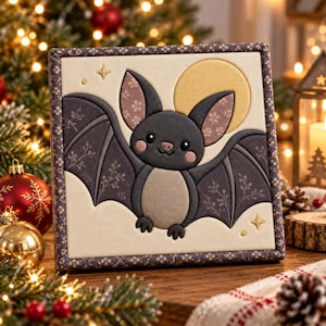 May include: A square fabric wall hanging featuring a central applique of a dark gray bat with large ears and pink cheeks, set against a cream background with a yellow moon and small gold stars, framed by a dark patterned fabric border.