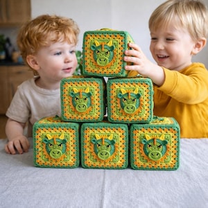 Może przedstawiać: Stos sześciu szydełkowych klocków do zabawy. Każdy klocek ma zieloną twarz smoka z żółtymi rogami i żółto-pomarańczową obwódką. Klocki są zielone, żółte i pomarańczowe. Klocki są ułożone na szarej powierzchni.