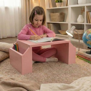 Puede incluir: Un escritorio portátil de madera rosa con almacenamiento de lápices es utilizado por una persona que escribe en un cuaderno. Un globo terráqueo, libros y una lámpara de escritorio están cerca. El escritorio está sobre una alfombra beige.