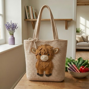 May include: A beige crocheted tote bag with a brown felt Highland cow applique. The bag has two handles and a bow detail. A bowl of vegetables sits nearby. The bag is on a wooden surface.