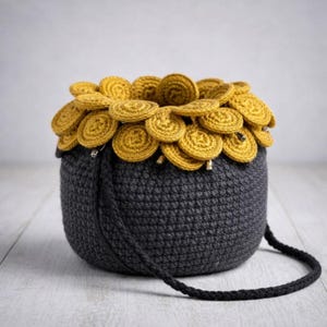 Może przedstawiać: Ręcznie robiona torba z szydełka w kolorze antracytowej szarości, z ozdobnym obramowaniem z okrągłych, złotożółtych akcentów. Torba ma pasujący czarny pasek i jest zaprojektowana w zaokrąglonym, cylindrycznym kształcie. Unikalny dodatek.