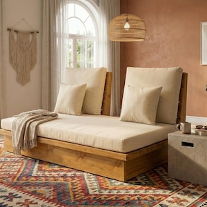 May include: A wooden daybed with cream-colored cushions and pillows sits on a colorful patterned rug. A woven pendant light hangs above. A small concrete side table holds a mug and sunglasses. A macrame wall hanging is visible.