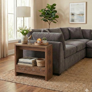 Może przedstawiać: Salon z ciemnoszarą sofą narożną, drewnianym stolikiem bocznym i dywanem jutowym. Stolik boczny ma dolną półkę z książkami i wiklinowym koszem. Wazon z białymi kwiatami i zapalona świeca stoją na wierzchu.