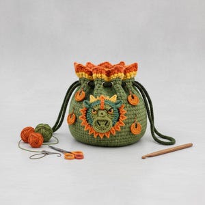 May include: A handmade green crochet drawstring bag featuring a dragon face applique. The bag has orange and yellow accents, a black drawstring, and small orange embellishments. Crochet hooks, scissors, and yarn balls are also visible.