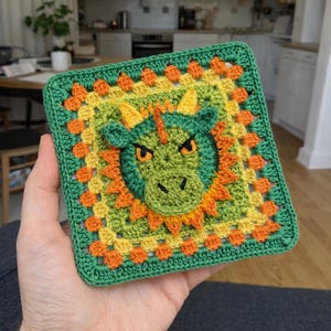 Peut inclure: Un visage de dragon carré, crocheté dans les tons de vert, jaune et orange. Le dragon a des cornes jaunes et une bande rouge sur la tête. Le carré est bordé de vert.