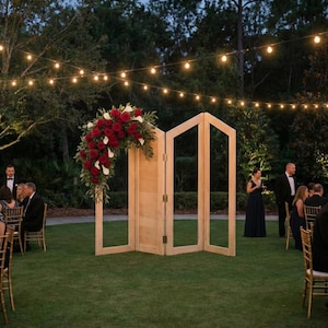 May include: A wooden folding screen with an arch of red roses and white flowers, set on a green lawn. The scene is lit by string lights, suggesting an outdoor event or wedding.